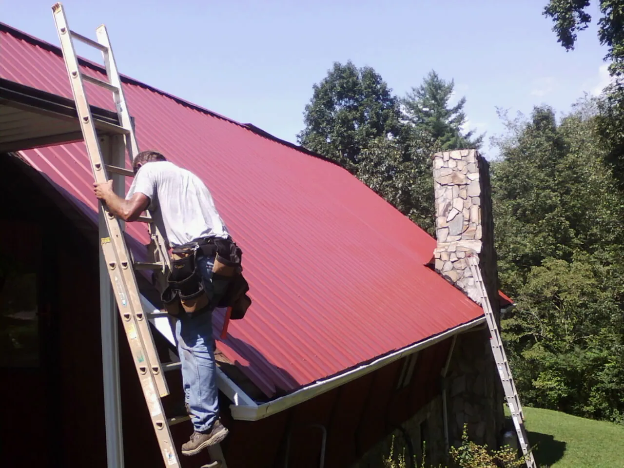 Roofer on a ladder beside a red metal roof and stone chimney — Soffit & Fascia Repair by Roofing Brothers in Lone Oak